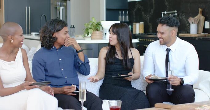 Diverse friends in formal wear celebrating laughing and talking after toasting on couch with plates