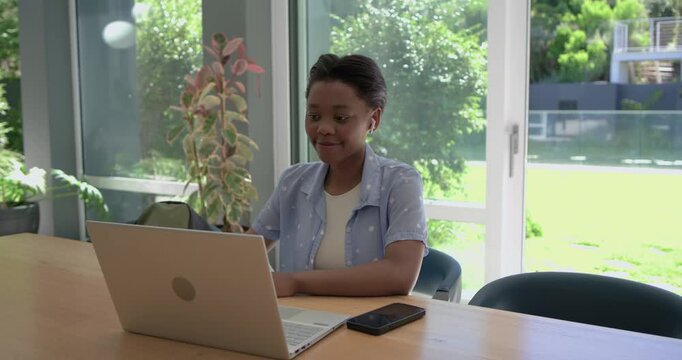 African American teenager sitting at home table earbuds hearing laptop ping cheering checking notes