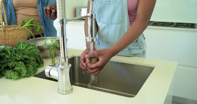 African mother and daughter washing roots under running faucet at kitchen sink, preparing meal