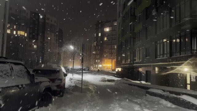 Modern buildings and cars under falling snow on city street at night. Camera moving