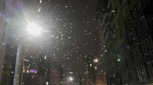 Modern buildings under falling snow on city street at night, low angle view. Camera moving