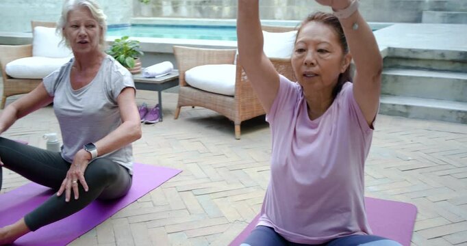 Diverse female friends stretching on purple mats at patio, following phone timer for flexibility