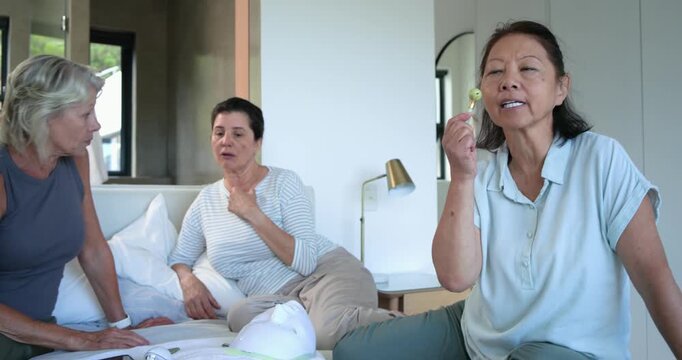 Diverse women sitting on bed, Asian senior trying green roller after friends pointing, copy space