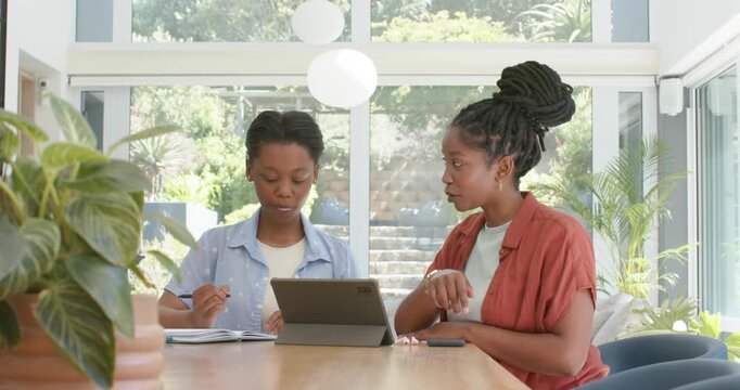 African American mom and teen daughter at table pointing at tablet daughter taking notes