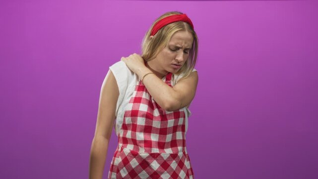 Woman chef in red check apron holding her shoulder and wincing while wearing red headband and watch in a studio; discomfort.