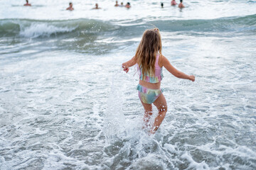 Fototapeta premium girl in colorful swimsuit jumping by sea shore, girl playing in the shallow water near seashore, waves warm summer day, family summer vacation kids splashing, enjoying carefree childhood play moments