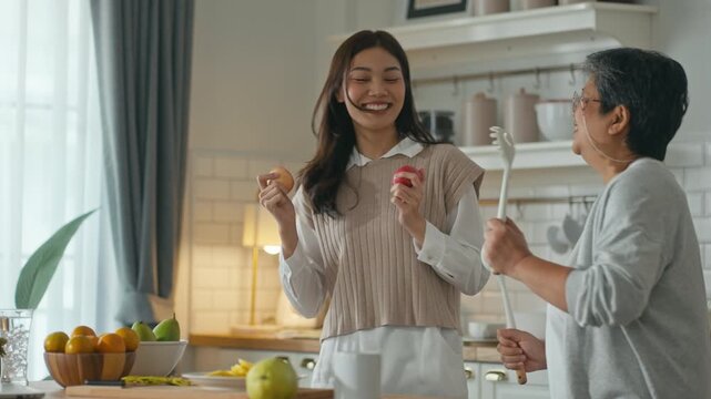 Happy senior Asian mother and adult daughter dancing and singing with kitchen utensils while cooking together at home. Concept of Mother's Day,family bonding,mom day and joyful domestic lifestyle.