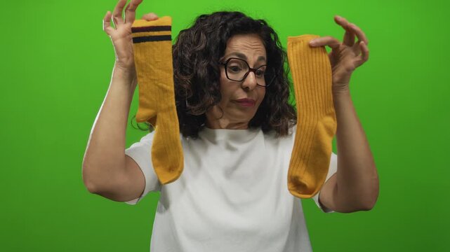 Middle age hispanic woman holds two different socks over a green studio wall; dilemma indecision uncertainty confusion.