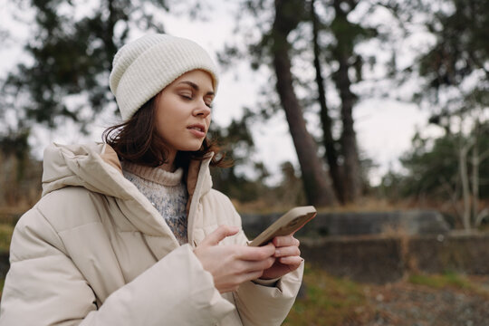 Woman in a warm coat using a smartphone outdoors in a park, winter light, calm moment, trees in background, cozy apparel and candid tech use
