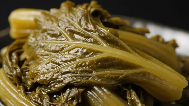 Extreme close-up shot of glossy, rich green Asian preserved mustard greens (Zha Cai or pickled cabbage) showing texture and leaf veins against a dark background.