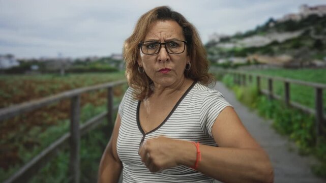 Woman wearing glasses extends hand forward through green field walkway lined by wooden fence rails; determination.