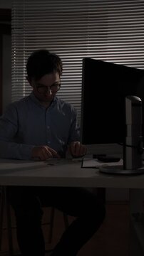 Vertical video man with glasses working late at desk, furtively slipping a small dark object into his shirt pocket near a dim monitor