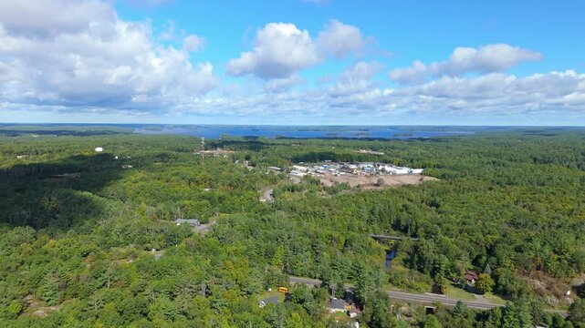 Aerial drone view of industrial buildings surrounded by dense forest with a lake visible in the distance near Gravenhurst, Muskoka, Ontario, showing rural industry within natural landscape