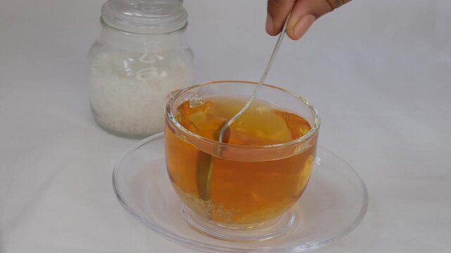 Hands of a woman preparing sweet tea, holding a spoon, pouring sugar powder into a glass mug of hot drink, adding sweetener or fructose to the beverage. Daily drink, unhealthy food concept. Closeup.