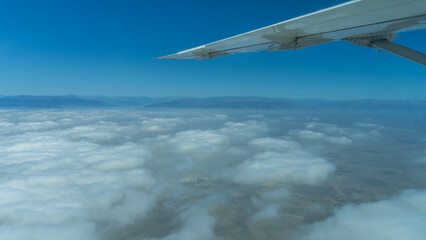Naklejka premium The wing of a flying airplane against the blue sky. Mountains on the horizon. Dense white cumulus clouds under the plane. The ground is visible through the gaps. Peru.