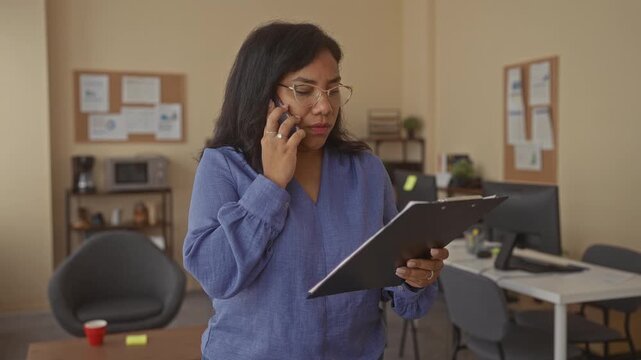 Woman in eyeglasses speaks on smartphone and holds clipboard with papers in modern office building; focus productivity organization efficiency.