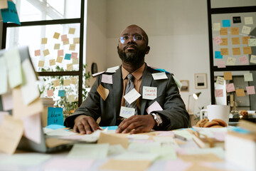 Black man sitting at cluttered office desk covered with sticky notes, wearing glasses and suit,...