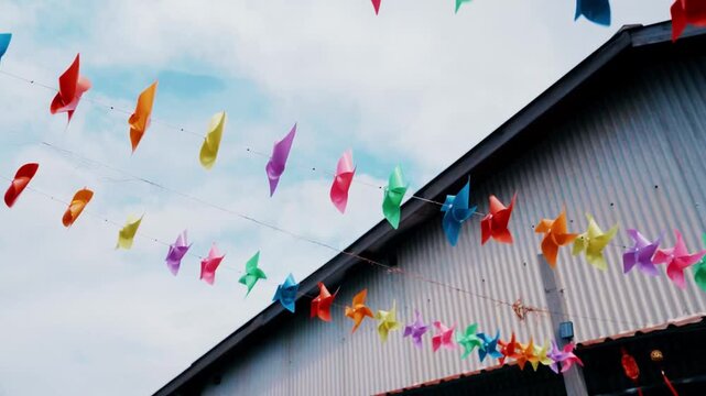 Rows of bright, multi-colored pinwheels or wind spinners hang on strings against the industrial texture of a grey building and a cloudy blue sky in Penang.