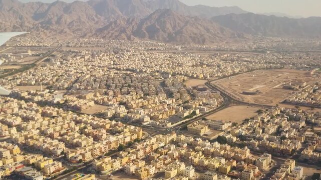 Aerial view from an airplane window over Aqaba, Jordan, showing a typical Middle Eastern desert settlement with dense buildings and dramatic mountain ranges on the horizon as the plane descends