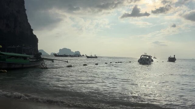 Long tail boats sit anchored near the shoreline at Railay Beach in Krabi, with calm sea water, floating buoys, distant islands and a cloudy sky creating a quiet coastal scene