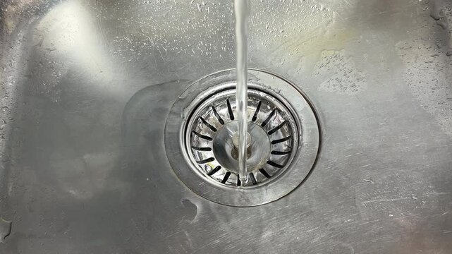 A medium-pressure stream of water flows from a tap into a stainless steel sink. The water hits the center of the metal drain, producing a constant and rhythmic splashing sound.