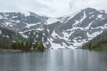 Mountain lake in rainy weather. Alpine lake between green hills with conifer forest with view to high rocky snowy mountain wall under gloomy cloudy sky. Circles of heavy rain on gray water surface.
