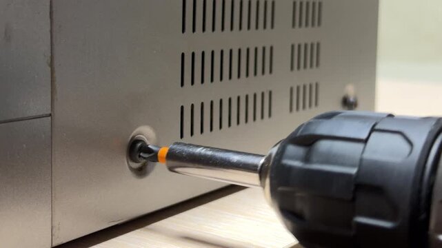 An electric screwdriver bit rotates rapidly to tighten a screw into a silver metal chassis. The friction between the metal components produces a harsh grinding sound and high-pitched mechanical whine.
