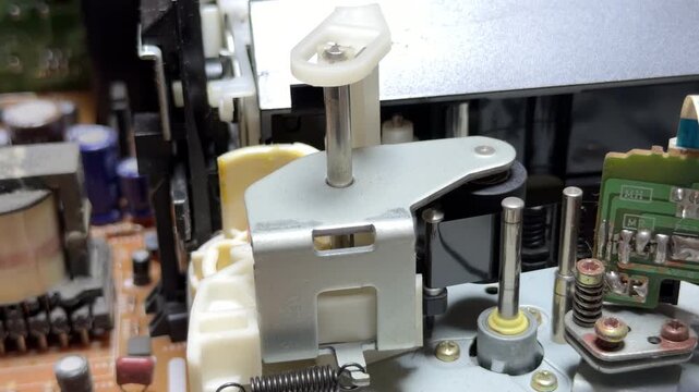 A vintage VCR mechanism pulls a magnetic tape into position against a rotating head. The internal gears and metal guides produce a rhythmic mechanical clatter and a steady whirring sound.