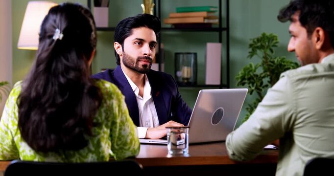 Indian couple visiting bank officer with laptop sitting across table during financial consultation discussing investment and real estate plans in modern corporate office interior setting