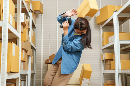 Asian adult woman small business owner shielding head while falling parcel boxes surround warehouse aisle, ecommerce storage error causing intense pressure during online seller daily work