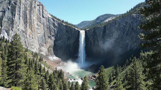 A majestic waterfall cascades down a rocky cliff face, surrounded by trees and mountains under a clear blue sky