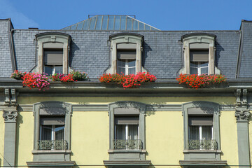 Classic European house facade with slate roof, dormer windows and colorful flower boxes under blue sky. Urban architecture exterior, travel concept. © Pavel