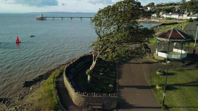 A drone shot of a band stand on the coast of Clevedon UK The shot plans to the left to reveal Clevedon Pier from behind some trees. It also shows Sailing boats then pans to reveal more coast
