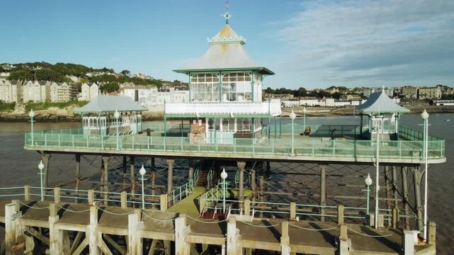 A drone shot face on to the end of Clevedon Pier UK with the coast in the background. The shot then rises up and forward towards the coast then tilts down to see the walkway up the pier