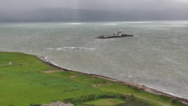 A cinematic aerial shot of Fenit Lighthouse on Little Samphire Island during a moody, overcast day. Perfect for travel documentaries and Irish coastal scenery.