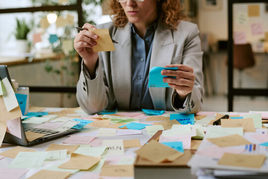 Caucasian woman sitting at desk organizing sticky notes and planning business tasks surrounded by laptop and paperwork in modern office workspace