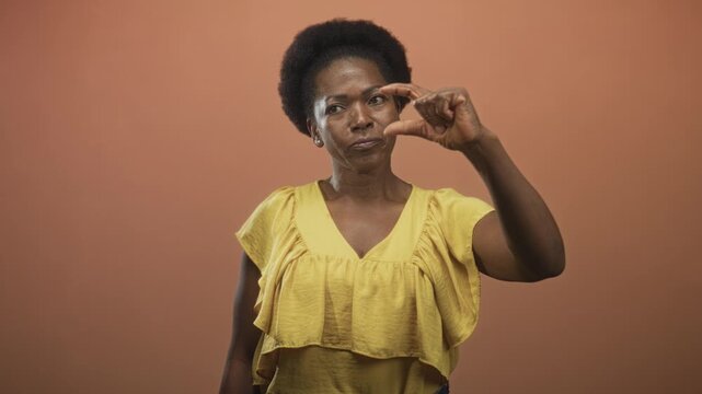Woman pinching fingers near eye while squinting and measuring a tiny gap, wearing yellow top against a studio backdrop; thoughtful focus.