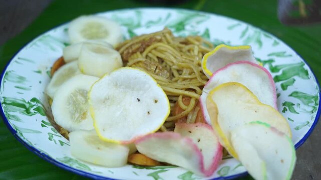 Delicious mie goreng served on a plate with sliced cucumbers and kerupuk or rice crackers on a green leaf background in Indonesia.