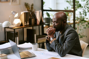 Black man wearing glasses sitting at desk praying in front of laptop screen, hands clasped in front of face, working in modern office environment with documents visible