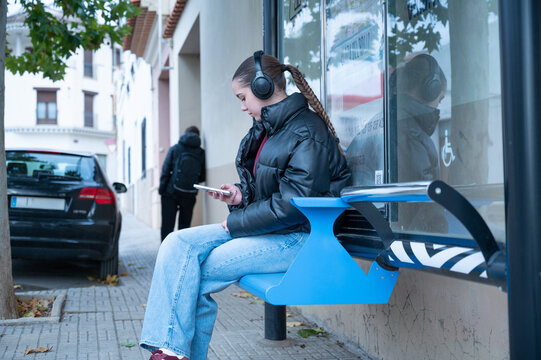 Teenage girl listening to music and checking smartphone at bus stop