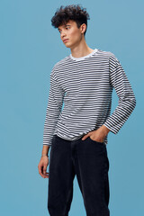 Young man wearing striped long sleeve shirt and dark pants posing against blue background, looking...