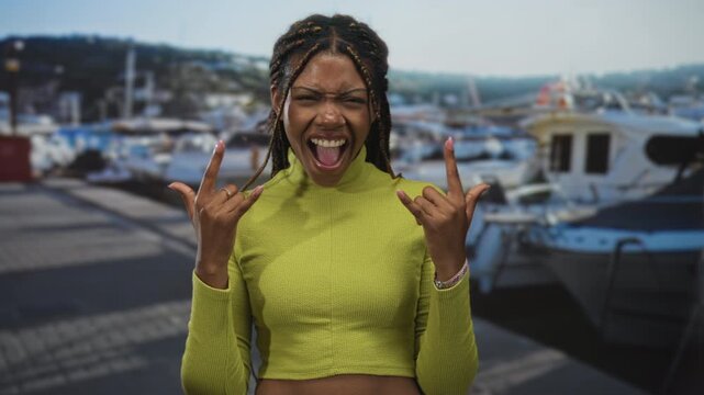 Woman laughing and flashing rock horns on a marina pier at a port with boats and docks; joy celebration fun.