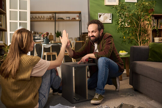 Caucasian young adult man and Caucasian young adult woman assembling furniture together in living room, smiling and giving high five while sitting on floor, teamwork and collaboration