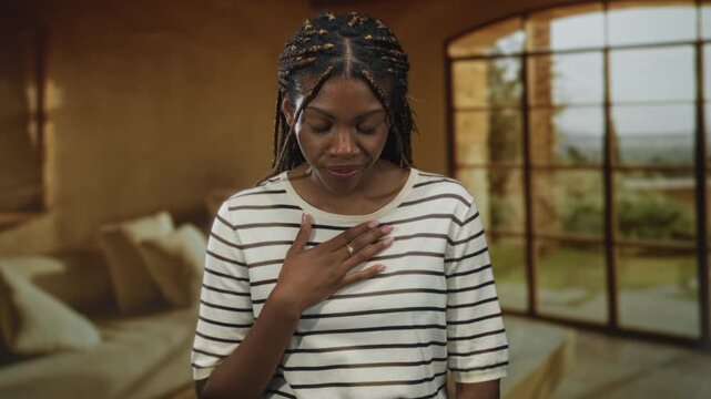 African american woman places hand on chest in studio living room set wearing striped top and braided hair visible; gratitude sincerity reflection.