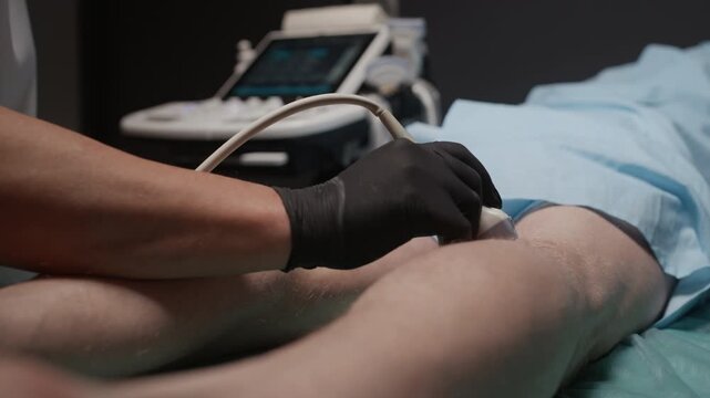 Cropped shot of gloved hands of sonographer moving ultrasound transducer while examining blood vessels of legs of unrecognizable patient lying on examination table in clinic