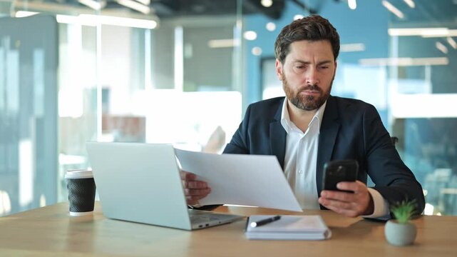 Professional man intently reviews documents, comparing information on smartphone and laptop. Demonstrates serious focus on business tasks and modern workplace efficiency, feeling concentrated.