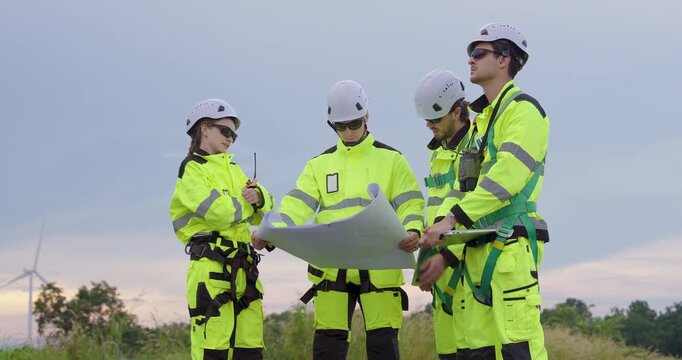 Caucasian engineer team in safety uniform discussing blueprint plan for renewable energy project near wind turbine, teamwork coordination maintenance schedule promoting sustainable power generation