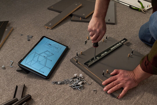 Caucasian middle aged man assembling furniture using screwdriver, following digital instructions displayed on tablet, hands working with screws and wooden boards on carpeted floor