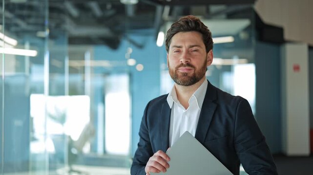 Professional stands within modern office, holding laptop, expressing confidence, happiness. Corporate success, positive future outlook conveyed through smile, posture. Ready for work.