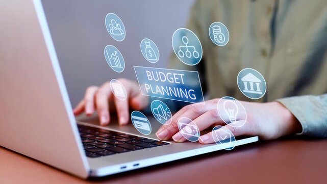 Hands typing on laptop with budget planning icons floating around on a table indoors in an office setting.